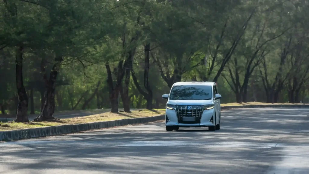 Toyota Alphard melaju di jalan beraspal yang teduh oleh pepohonan